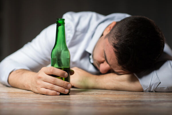 A man slumped over a table holding a beer bottle, illustrating how hard is it to stop drinking alcohol