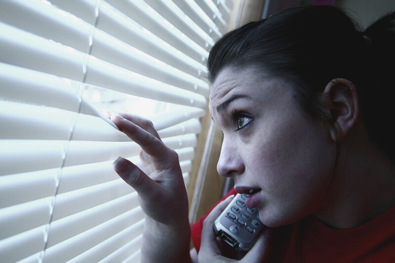  A young lady peeking out a window and on her phone, her expression reflecting paranoia, highlighting the importance of seeking support at a meth rehab in Phoenix, AZ