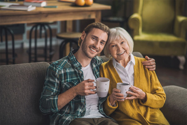 A young man and an elderly woman smiling and holding mugs on a couch, suggesting strong family support for addiction recovery.