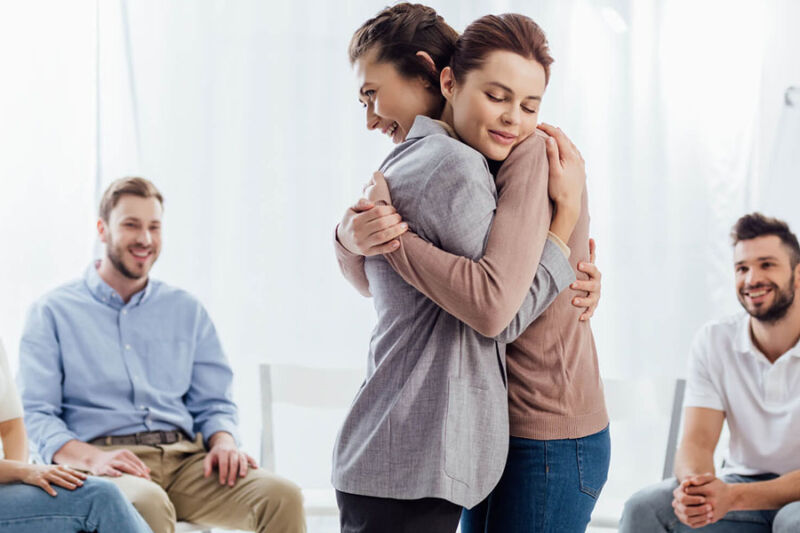 Two women embrace during a supportive group therapy session at a PHP Drug Rehab in Phoenix, with others smiling in the background.