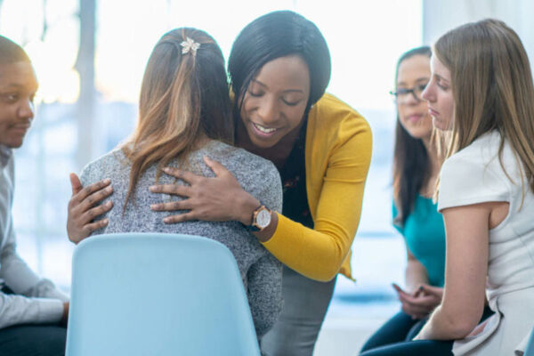 Young woman finding comfort and support in a group therapy session for mental health, embracing another participant with understanding and compassion.